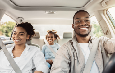 man and woman smiling in front seat of a car with little girl in the back smiling