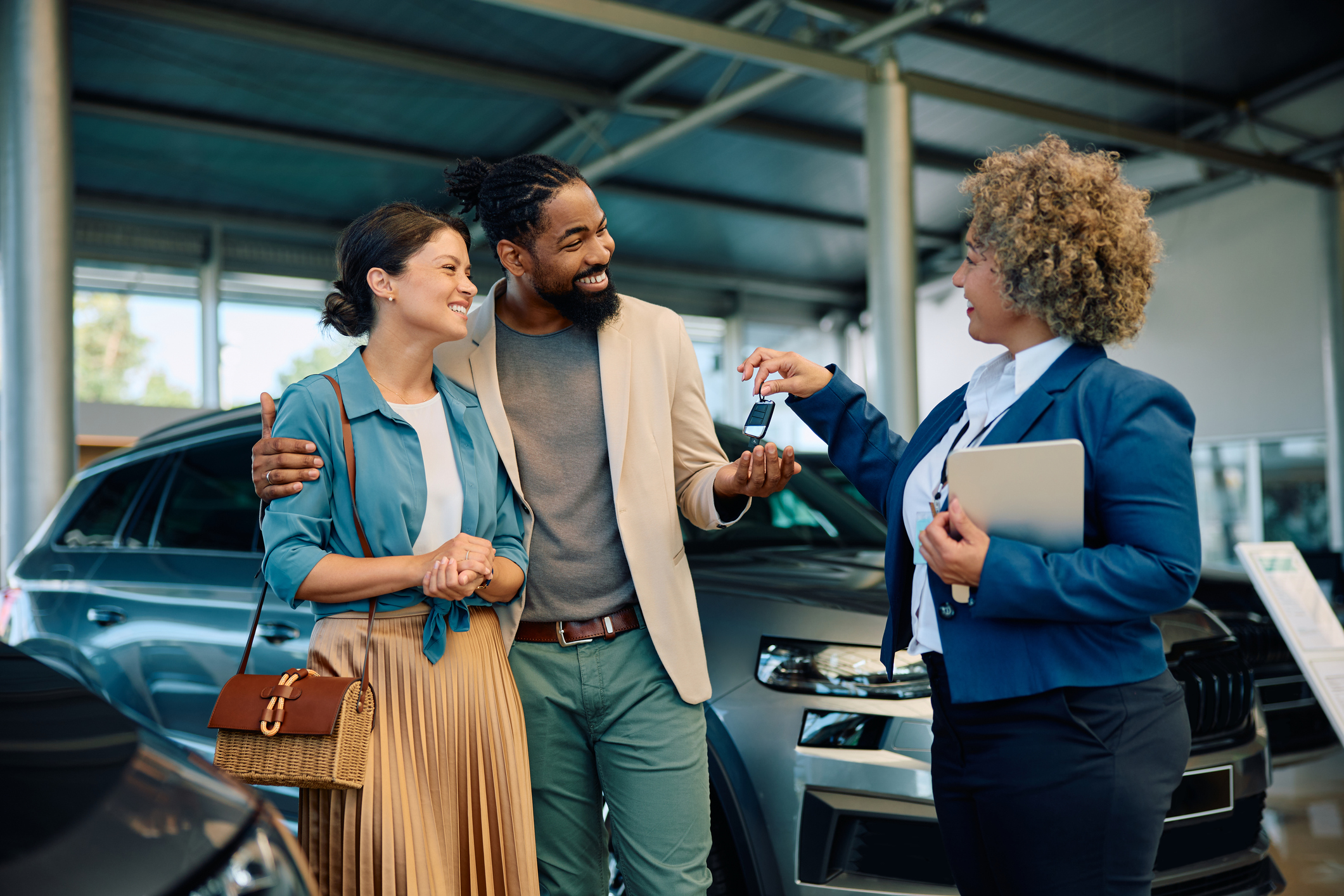 Happy multiracial couple receiving keys of their new car from saleswoman in showroom. 