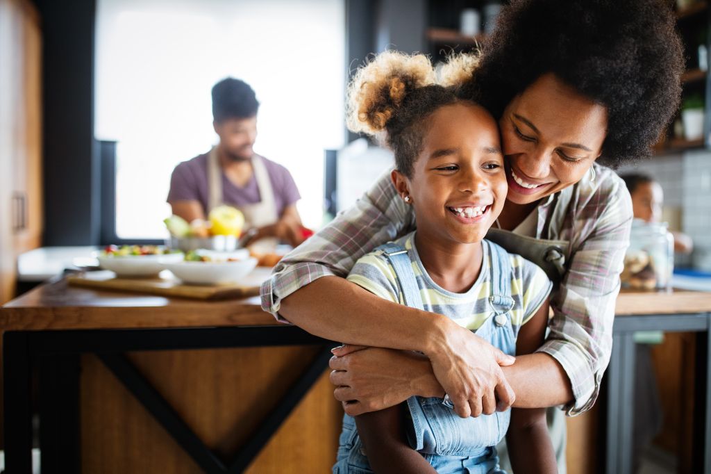 A smiling mother hugs her laughing daughter in a cozy kitchen, while another person prepares food in the background.
