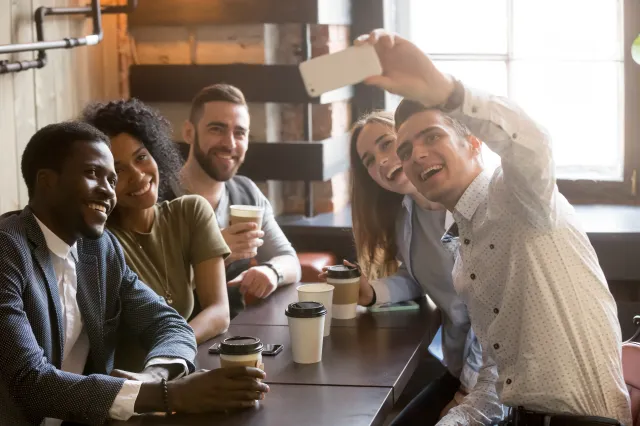 Un groupe de cinq jeunes adultes assis autour d’une table de café, souriants et prenant un selfie ensemble. Ils sont habillés de façon décontractée et semblent apprécier leur café et leur moment ensemble. L’endroit est lumineux grâce à la lumière naturelle qui entre par de grandes fenêtres.